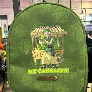 May include: Green backpack with a cartoon of a person holding a cabbage and a cart full of cabbages. The text on the backpack says "My Cabbages!" and "Handcrafted".