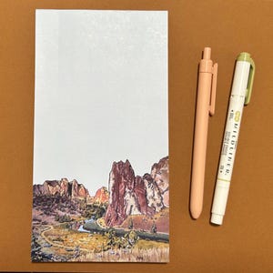 May include: A rectangular notepad featuring a landscape illustration of rocky cliffs and a winding river at the bottom, accompanied by a solid peach-colored retractable pen and a white Zebra Mildliner double-ended marker pen.