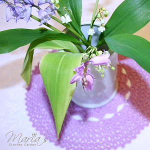 May include: A close-up of a bouquet of blue and white flowers in a white ceramic vase. The flowers are arranged on a purple crocheted doily. The text "Maria's Crochet Garden" is visible in the bottom left corner of the image.