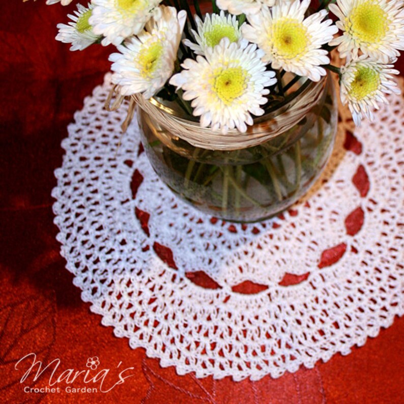 May include: White crochet doily with a delicate pattern, featuring a vase of white daisies on a red background. The doily is a circular design with a center hole and a scalloped edge.