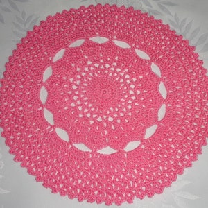 May include: A pink crocheted doily with a delicate floral pattern. The doily is circular and has a scalloped edge.