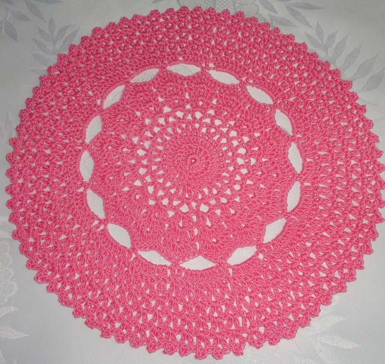 May include: A pink crocheted doily with a delicate floral pattern. The doily is circular and has a scalloped edge.