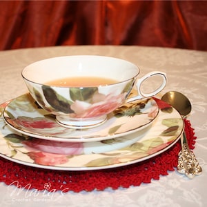 May include: A white teacup and saucer set with a floral design. The cup is filled with tea and the saucer has a matching floral pattern. The set is on a red crocheted doily.