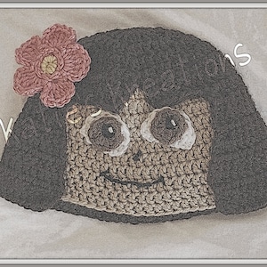 Puede incluir: Cabeza de muñeca de crochet con cabello castaño, ojos marrones y una flor rosa en la parte superior. La muñeca sonríe.