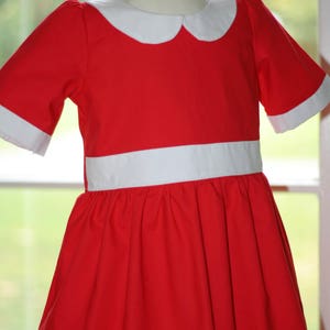 Op de afbeelding: Een rode jurk met een witte kraag en een witte riem. De jurk heeft een volle rok en korte mouwen. De jurk is gemaakt van een zachte, comfortabele stof. "Merri Poppins Children's Boutique" is gedrukt op de onderkant van de jurk.