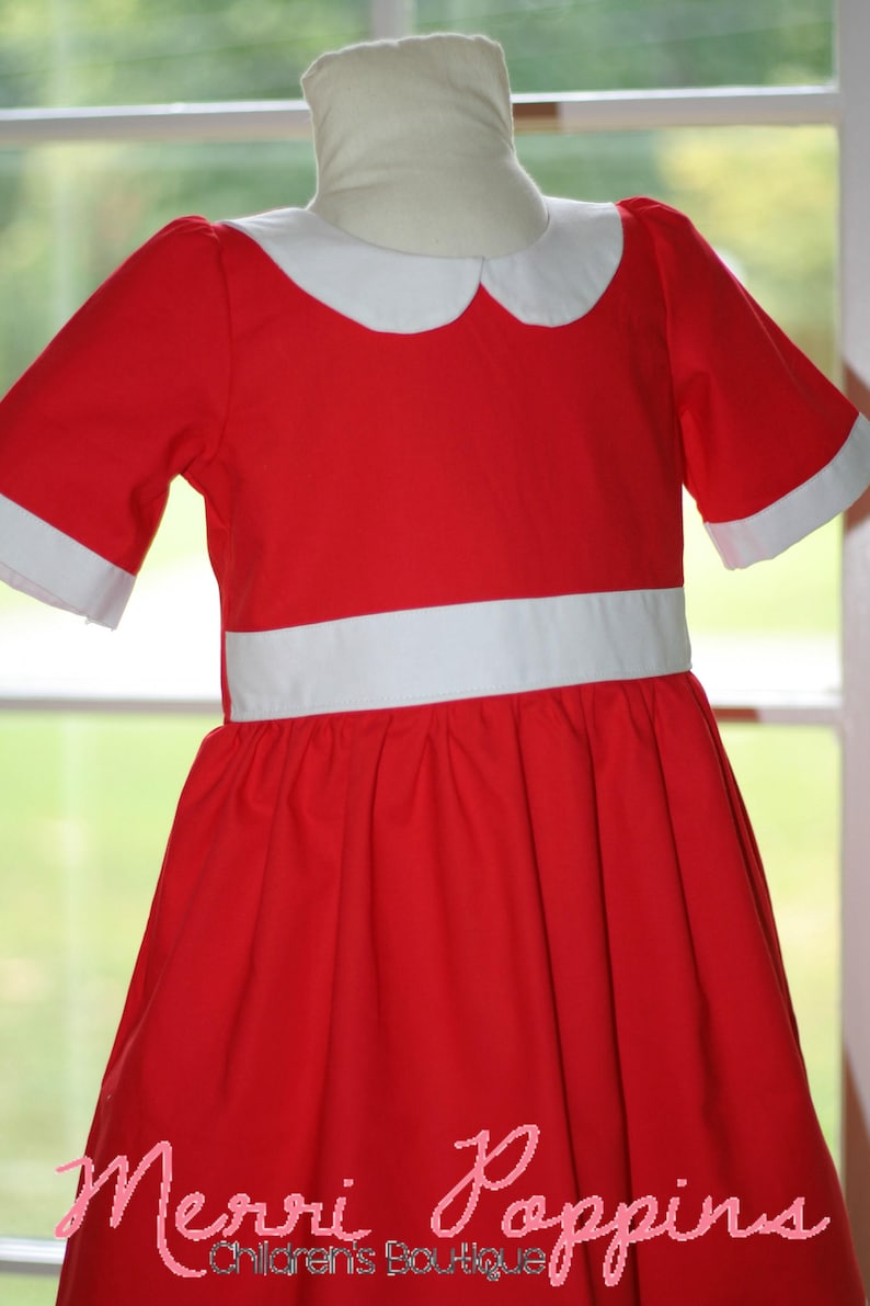 Op de afbeelding: Een rode jurk met een witte kraag en een witte riem. De jurk heeft een volle rok en korte mouwen. De jurk is gemaakt van een zachte, comfortabele stof. "Merri Poppins Children's Boutique" is gedrukt op de onderkant van de jurk.