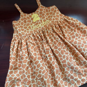 K&ouml;nnte beinhalten: Ein &auml;rmelloses Kleid mit braunem und wei&szlig;em Giraffenmuster, auf dem vorne eine gelbe Zahl "2" und der Name "Thunder" gestickt sind.