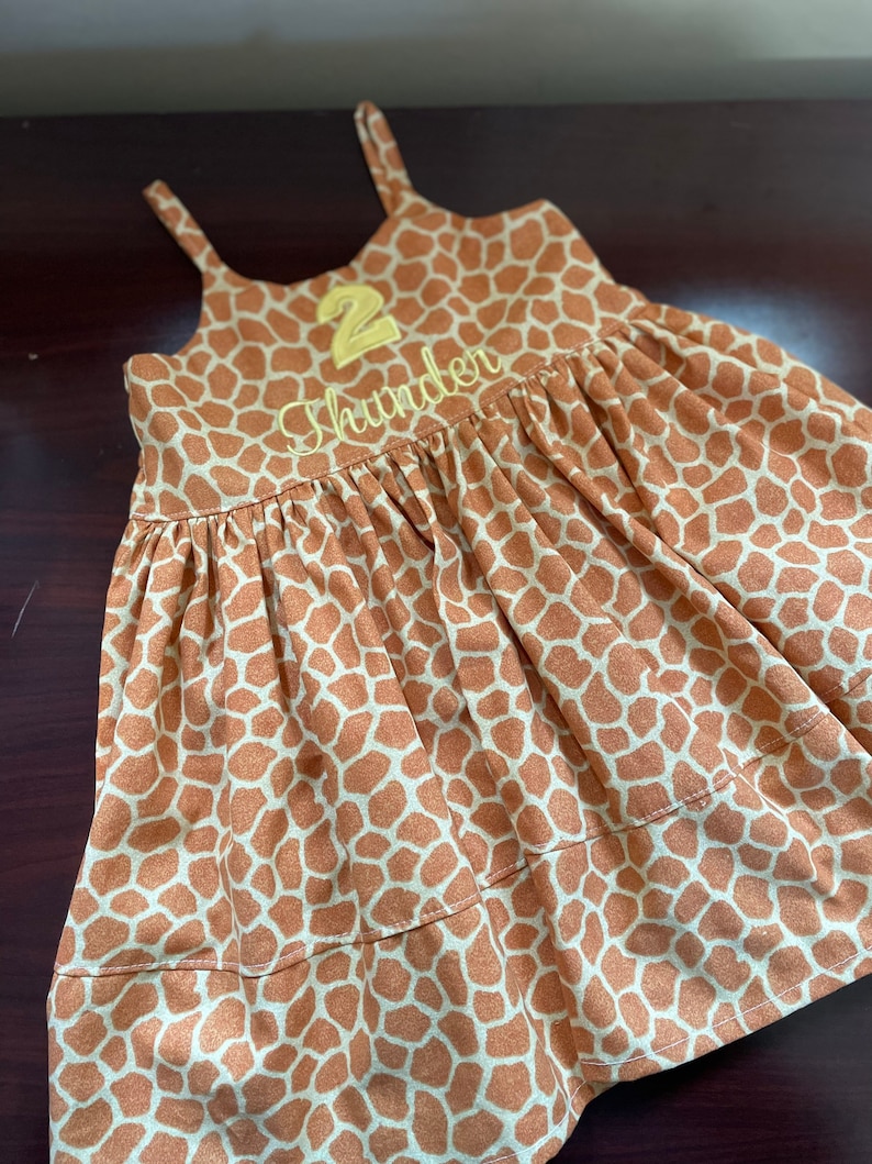 K&ouml;nnte beinhalten: Ein &auml;rmelloses Kleid mit braunem und wei&szlig;em Giraffenmuster, auf dem vorne eine gelbe Zahl "2" und der Name "Thunder" gestickt sind.