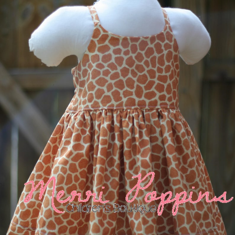 K&ouml;nnte beinhalten: Ein &auml;rmelloses Kleid mit Giraffenmuster in Braun- und Wei&szlig;t&ouml;nen. Das Kleid hat einen gerafften Rock und einen schlichten Oberteil. Das Kleid ist mit "Merri Poppins Children's Boutique" beschriftet.