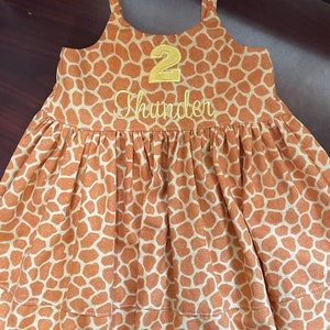 K&ouml;nnte beinhalten: Ein &auml;rmelloses Kleid mit braunem und wei&szlig;em Giraffenmuster, auf dem vorne eine gelbe Zahl "2" und der Name "Thunder" gestickt sind.