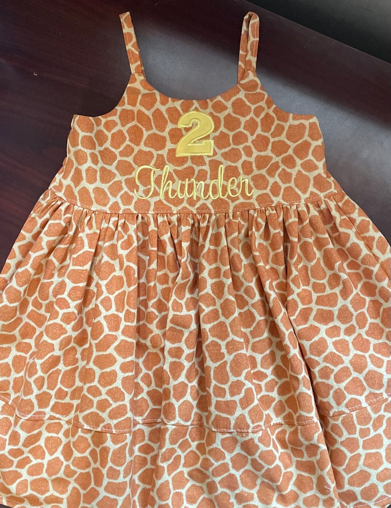 K&ouml;nnte beinhalten: Ein &auml;rmelloses Kleid mit braunem und wei&szlig;em Giraffenmuster, auf dem vorne eine gelbe Zahl "2" und der Name "Thunder" gestickt sind.