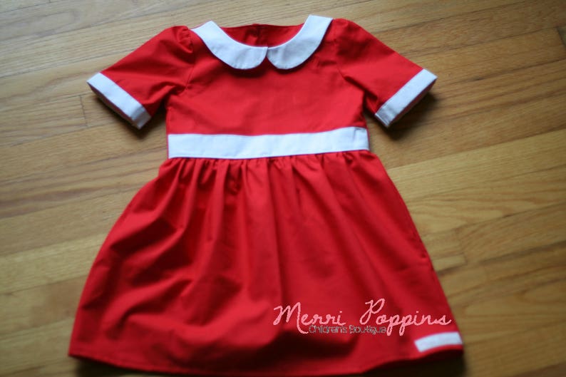 Op de afbeelding: Een rode jurk met een witte kraag en witte bies rond de taille. De jurk heeft een geplooide rok en is gelabeld "Merri Poppins Children's Boutique."