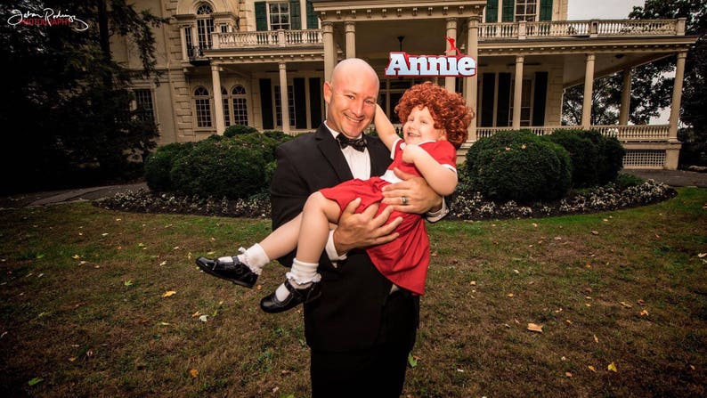 Op de afbeelding: Een man in een zwart smoking houdt een jong meisje vast dat is gekleed als Annie in een rode jurk. Het meisje glimlacht en kijkt naar de camera. De man glimlacht ook en kijkt naar de camera. De achtergrond is een groot, oud huis met een bord waarop "Annie" staat.
