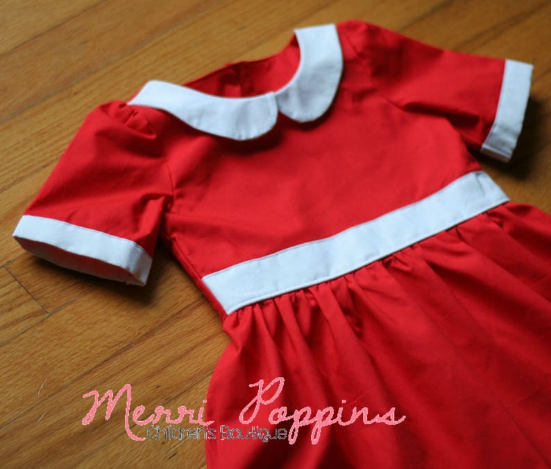 Op de afbeelding: Een rode jurk met witte bies en een witte Peter Pan-kraag. De jurk heeft een geplooide rok en korte mouwen. De tekst "Merri Poppins Children's Boutique" is zichtbaar onderaan de afbeelding.