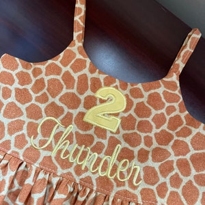 K&ouml;nnte beinhalten: Ein braunes und hellbraunes, &auml;rmelloses Kleid mit Giraffenmuster. Das Kleid hat eine gelbe Nummer "2" und den Namen "Thunder" vorne aufgestickt.