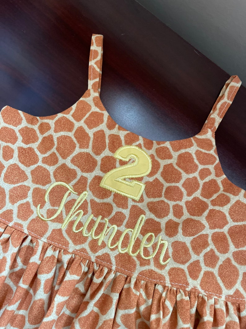 K&ouml;nnte beinhalten: Ein braunes und hellbraunes, &auml;rmelloses Kleid mit Giraffenmuster. Das Kleid hat eine gelbe Nummer "2" und den Namen "Thunder" vorne aufgestickt.