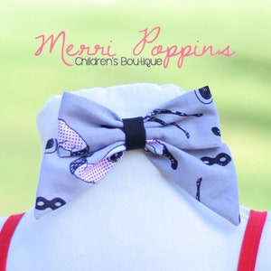 May include: A grey bow tie with a black band, decorated with black and purple mask and eye patterns. The bow tie is shown on a white surface with red braces visible. Text reads "Merri Poppins Children's Boutique."