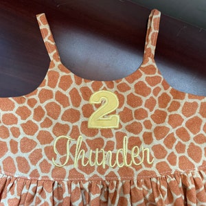 K&ouml;nnte beinhalten: Ein braunes und hellbraunes, &auml;rmelloses Kleid mit Giraffenmuster. Das Kleid hat eine gelbe Nummer "2" und den Namen "Thunder" auf der Vorderseite gestickt.