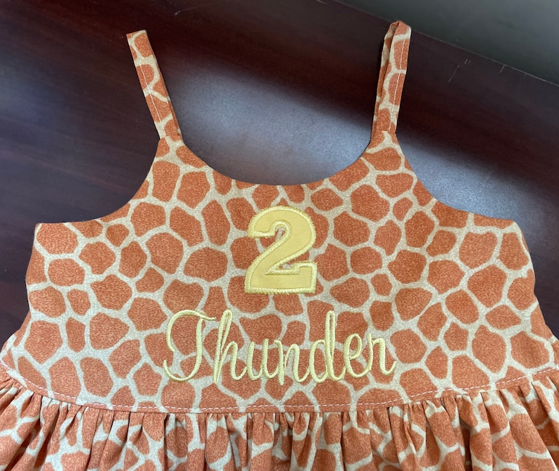 K&ouml;nnte beinhalten: Ein braunes und hellbraunes, &auml;rmelloses Kleid mit Giraffenmuster. Das Kleid hat eine gelbe Nummer "2" und den Namen "Thunder" auf der Vorderseite gestickt.