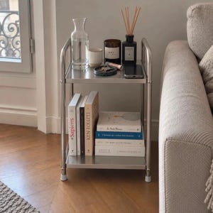 May include: A two-tier stainless steel rolling cart with a top shelf holding a clear glass carafe, a small ceramic cup, a candle labeled A.P.C., a reed diffuser, a small tray with keys, and a smartphone. The bottom shelf holds five stacked books.