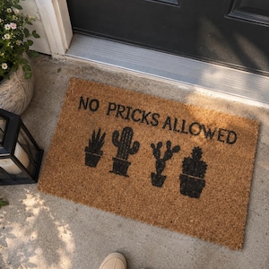 Puede incluir: Un felpudo rectangular de fibra de coco beige con el texto negro NO PRICKS ALLOWED sobre cuatro siluetas negras de diferentes cactus en macetas. La alfombra está colocada sobre una superficie de hormigón frente a una puerta oscura.