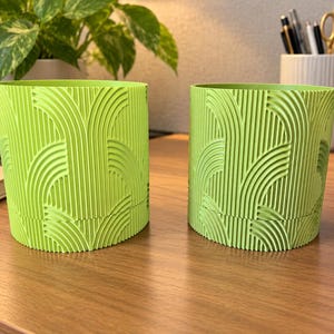 May include: Two cylindrical lime green planters featuring a raised, textured pattern of vertical lines and repeating semi-circular arches. Each planter has a distinct horizontal seam near the base, indicating a two-part construction.
