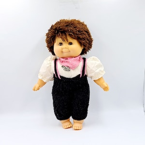 May include: A brown haired doll with a pink polka dot bandana and black overalls. The doll is wearing a white shirt with a silver charm.