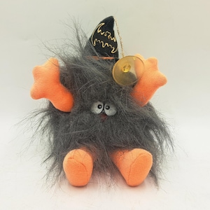 May include: A plush toy featuring a fuzzy dark gray body with bright orange hands and feet. The creature wears a black pointed hat decorated with a gold embroidered bat design and includes a clear plastic suction cup attached by a gold cord.