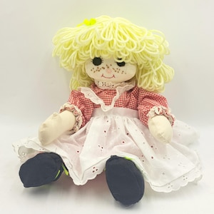 May include: A handmade rag doll with yellow yarn hair and freckles. The doll wears a red and white gingham dress with a white eyelet apron and black shoes. A yellow flower is pinned in the hair. The doll is a vintage toy.