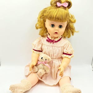 May include: A vintage doll with blonde hair, wearing a pink and white striped dress and white socks, holding a small teddy bear.