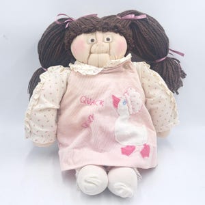 May include: A vintage doll with brown yarn hair styled in pigtails, tied with pink ribbons. The doll wears a pink corduroy dress with a white duck applique and the word "QUACK" embroidered on it. The doll has rosy cheeks and white socks.