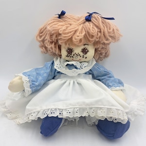 May include: A handmade rag doll with a light peach yarn hairstyle, blue bows, and a painted face with freckles. The doll wears a blue floral print dress with a white eyelet lace collar and apron, and blue shoes. A vintage-style toy.