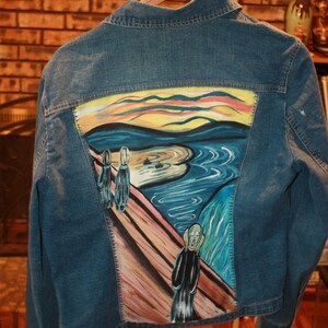 May include: A blue denim jacket with a painting of The Scream by Edvard Munch on the back. The painting is in the style of the original, with a yellow sky, a swirling blue water, and a figure with a screaming face.