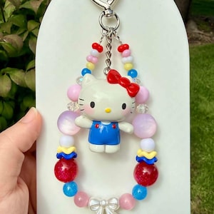 May include: A keychain featuring a Hello Kitty figure wearing a blue outfit and a red bow, suspended from a heart-shaped metal clasp. The figure is surrounded by a loop of colorful beads in shades of red, blue, pink, yellow, and white, with a white bow charm at the bottom.