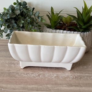 May include: A rectangular off-white ceramic planter with a scalloped, fluted exterior design. The vessel features a smooth, glossy finish and is supported by four small, integrated feet at the base, providing a stable foundation for indoor plants.