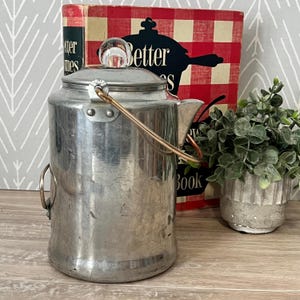May include: A vintage metal tea kettle with a clear glass knob on the lid and a copper-colored handle. The kettle is cylindrical with a spout and a small handle on the side. A cookbook with the title "Better Homes" is in the background.