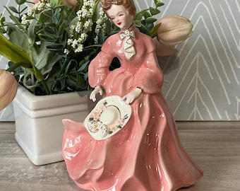 May include: A vintage porcelain figurine of a woman in a long, flowing pink dress, holding a hat with floral accents. The figurine has a delicate face and is posed gracefully. The hat features a floral design. The figurine is a decorative item.