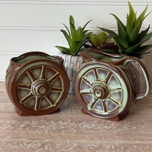May include: Two brown and green ceramic vessels, one a rounded planter and the other a pitcher, both featuring a raised wagon wheel design on the front. The glaze is a deep brown with lighter green accents highlighting the wheel spokes and rims.