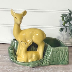 May include: A ceramic planter featuring a yellow deer and fawn figurine set within a green, tree-stump-shaped base. The planter has a rectangular opening for plants. The deer are a light yellow color.