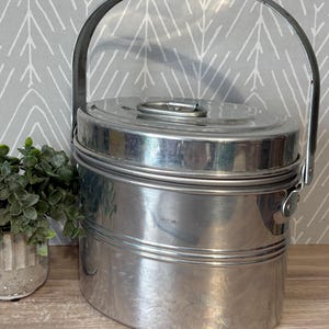 May include: A vintage, silver-colored metal lunch pail with a rounded rectangular shape. It features a hinged lid with a circular handle and a sturdy, arched handle for carrying. The lunch pail has horizontal bands and a latch on the side.