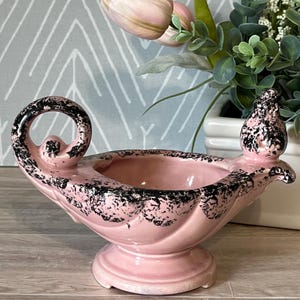 May include: A pink ceramic decorative bowl with a pedestal base and a handle. The bowl has a scalloped rim and is accented with black speckles. The bowl is likely vintage and is suitable for display.