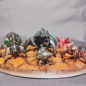 May include: A collection of six miniature insectoid creatures with varied dark, green, and red colorations, positioned on a textured, cracked orange-brown base that mimics a dry, arid landscape with uneven terrain and rocky details.