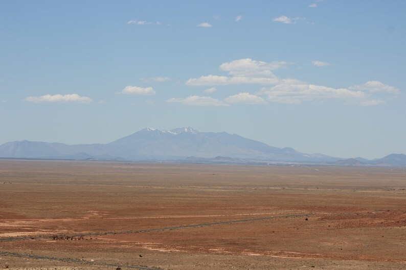 May include: A vast, arid desert landscape with reddish-brown earth in the foreground, stretching toward a distant, hazy mountain range under a clear blue sky with scattered white clouds. The peaks show faint traces of white snow.