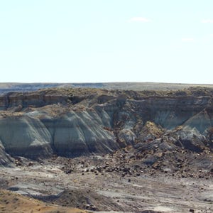Puede incluir: Un vasto paisaje des&eacute;rtico con formaciones de roca sedimentaria en capas en tonos bronceado, gris y marr&oacute;n oscuro. El terreno se caracteriza por colinas erosionadas y barrancos bajo un cielo azul claro con escasa vegetaci&oacute;n.