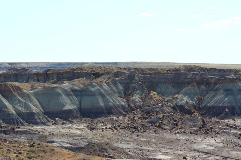 Puede incluir: Un vasto paisaje des&eacute;rtico con formaciones de roca sedimentaria en capas en tonos bronceado, gris y marr&oacute;n oscuro. El terreno se caracteriza por colinas erosionadas y barrancos bajo un cielo azul claro con escasa vegetaci&oacute;n.