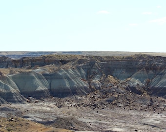 May include: A vast desert landscape featuring layered sedimentary rock formations in shades of tan, gray, and deep brown. The terrain is characterized by eroded hills and gullies under a clear, bright blue sky with sparse vegetation.