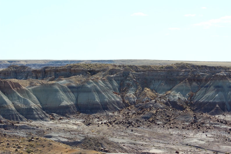 May include: A vast desert landscape featuring layered sedimentary rock formations in shades of tan, gray, and deep brown. The terrain is characterized by eroded hills and gullies under a clear, bright blue sky with sparse vegetation.