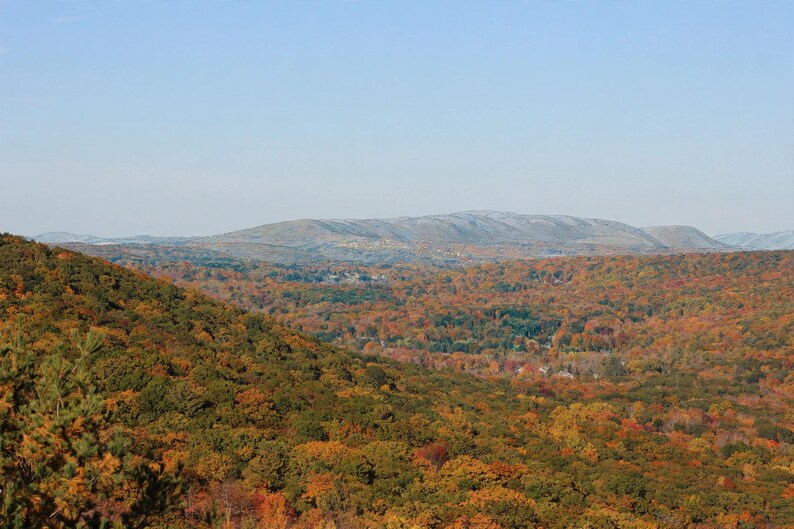 K&ouml;nnte beinhalten: Eine weite Landschaftsaufnahme eines dichten Waldes im Herbst mit einem leuchtenden Bl&auml;tterdach in Orange, Rot, Gelb und Gr&uuml;n. In der Ferne erheben sich h&uuml;gelige Berge vor einem klaren, hellblauen Himmel bei hellem Tageslicht.