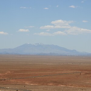 Puede incluir: Un vasto paisaje des&eacute;rtico &aacute;rido con tierra de color marr&oacute;n rojizo en primer plano, que se extiende hacia una cadena monta&ntilde;osa lejana y brumosa bajo un cielo azul claro con nubes blancas dispersas. Los picos muestran rastros de nieve.