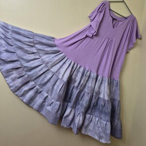 May include: A flowing, lavender dress with short sleeves and a V-neck. The bodice has a perforated design, and the skirt features layered ruffles in shades of lavender and gray. The dress is hanging on a metal hanger.